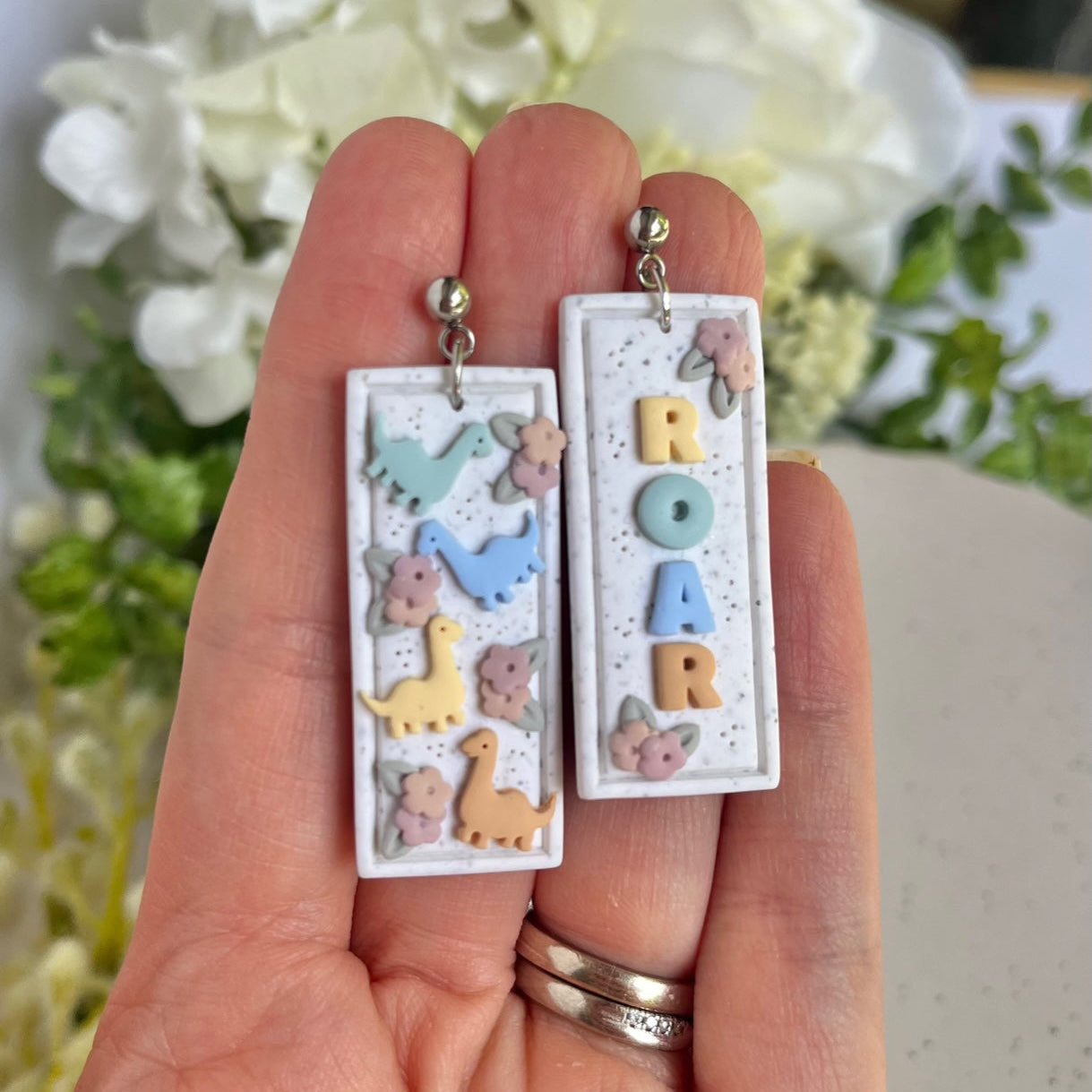a set of earrings with 'ROAR' and dinosaur designs against a floral background