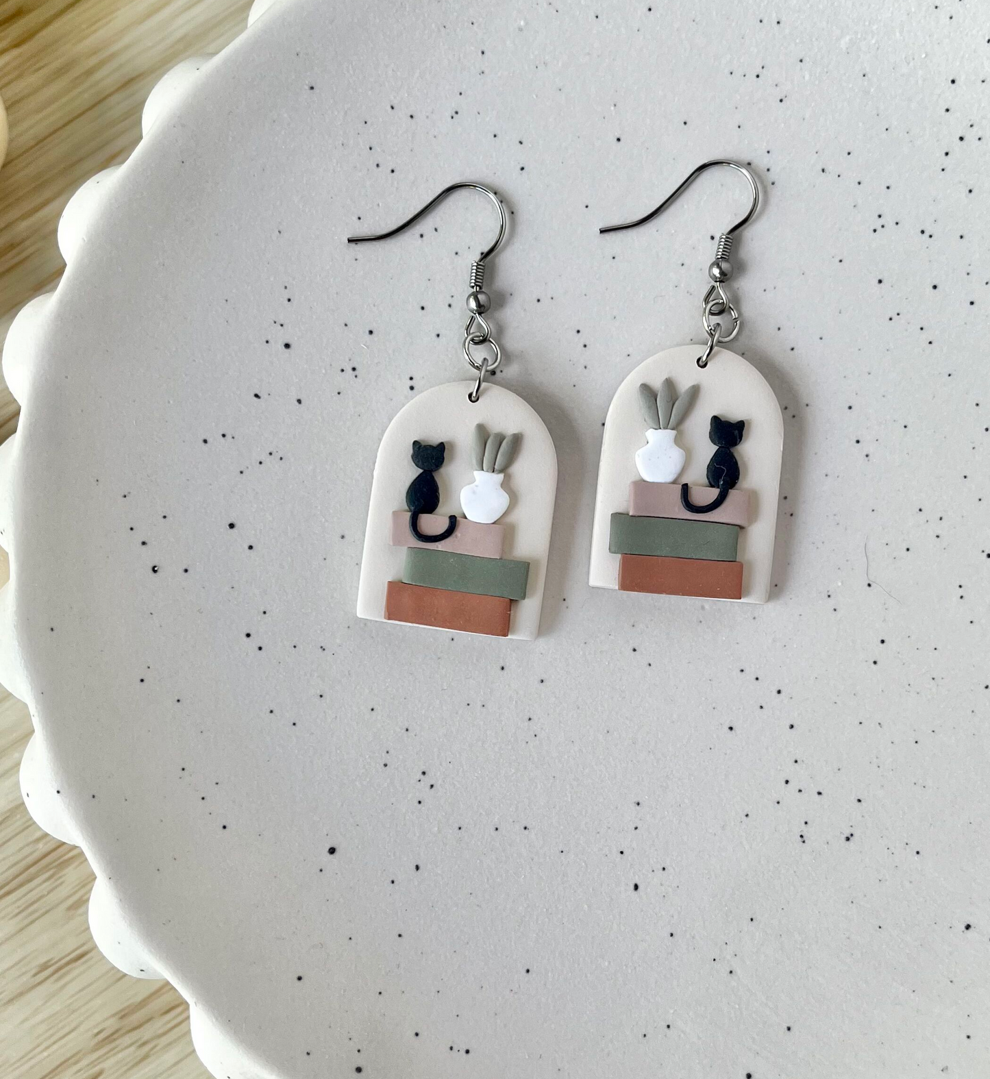 A pair of earrings featuring a black cat atop a stack of books with a tiny potted plant, displayed on a white plate.