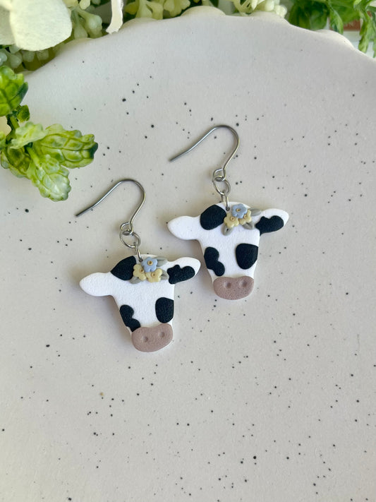 Cow-shaped earrings on a white surface with greenery in the background