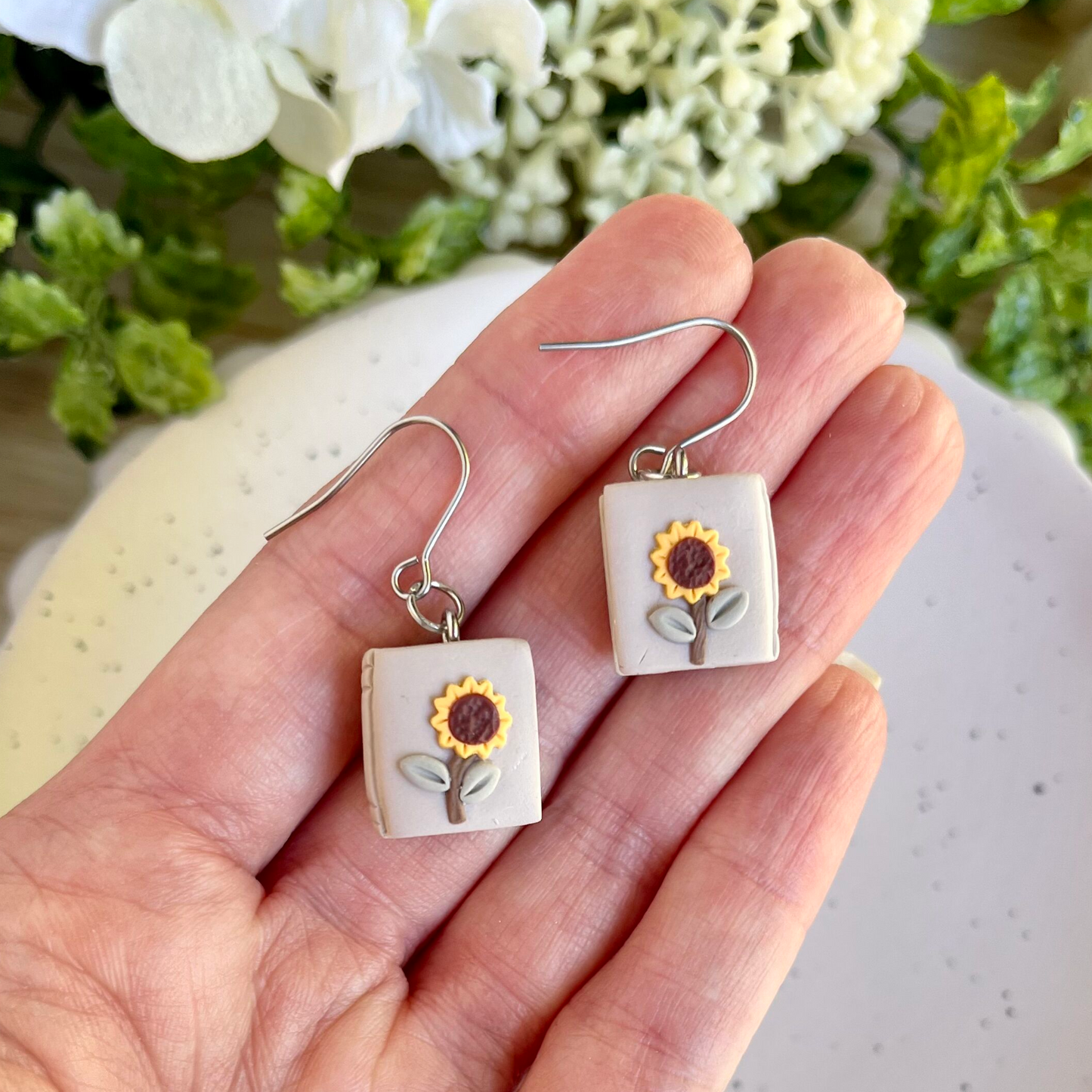 A pair of square-shaped earrings with sunflower designs, held in a hand against a floral backdrop.