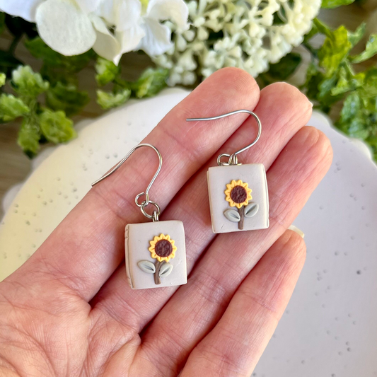 A pair of square-shaped earrings with sunflower designs, held in a hand against a floral backdrop.