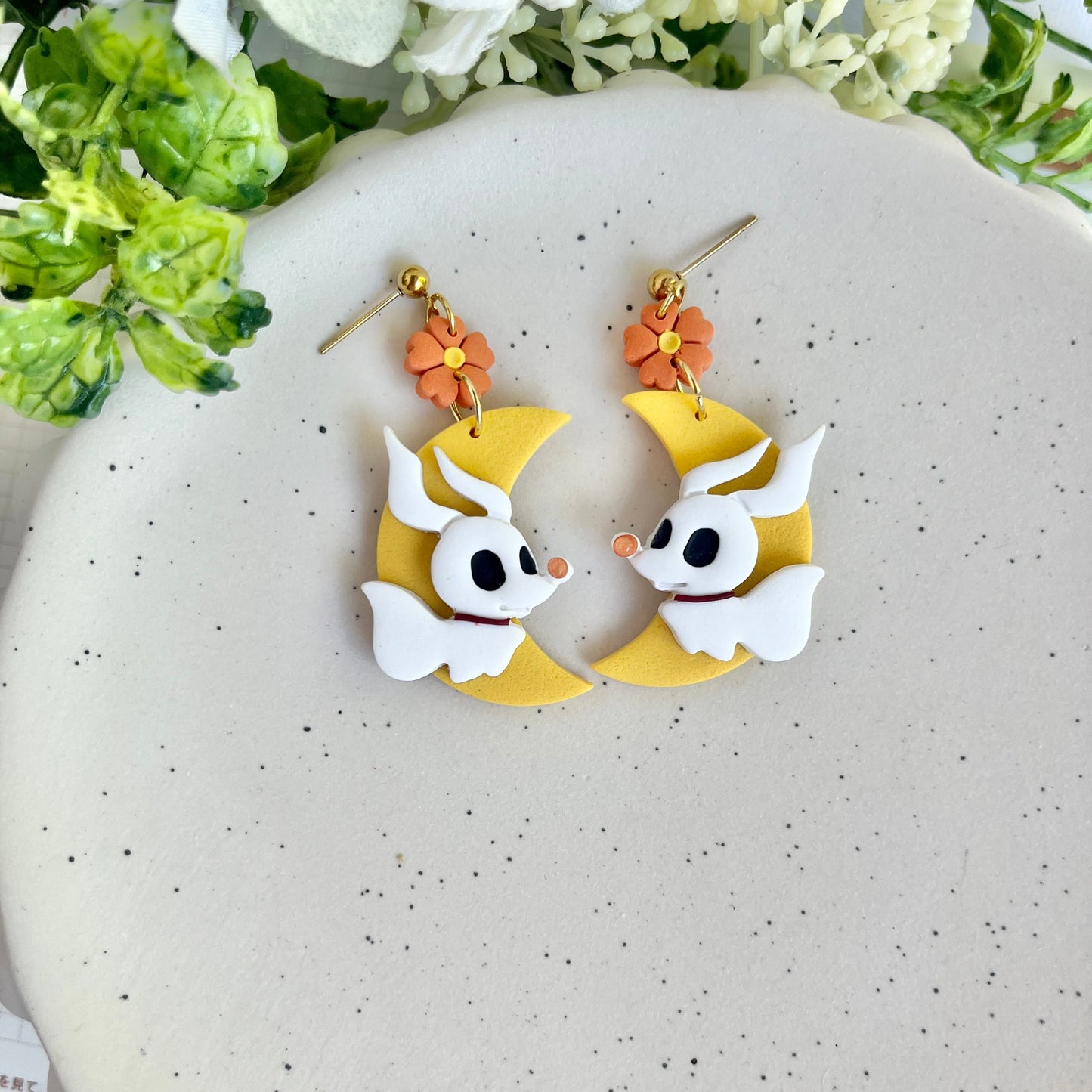 Pair of earrings with cartoon character design on a speckled surface with flowers in the background