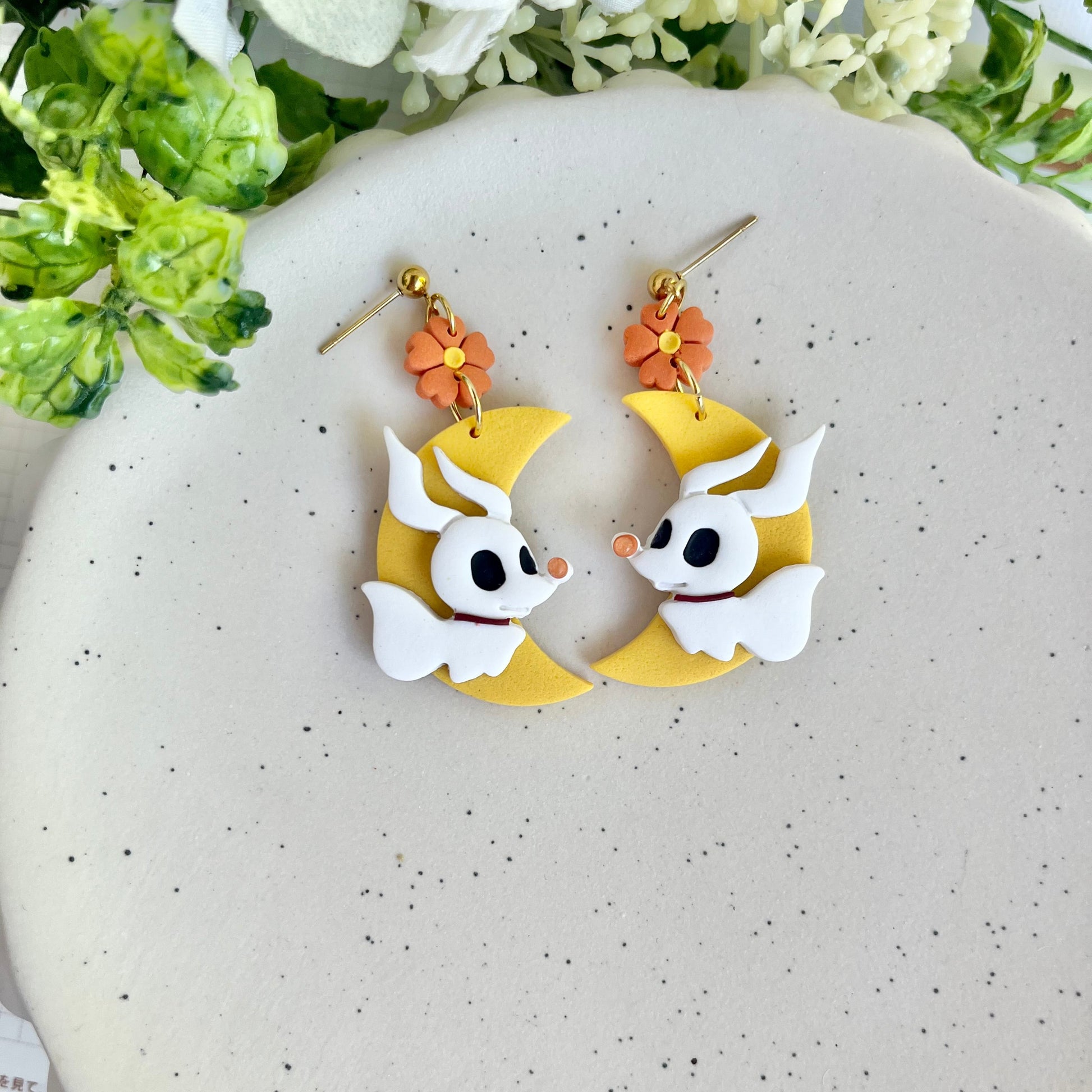 Pair of earrings with cartoon character design on a speckled surface with flowers in the background