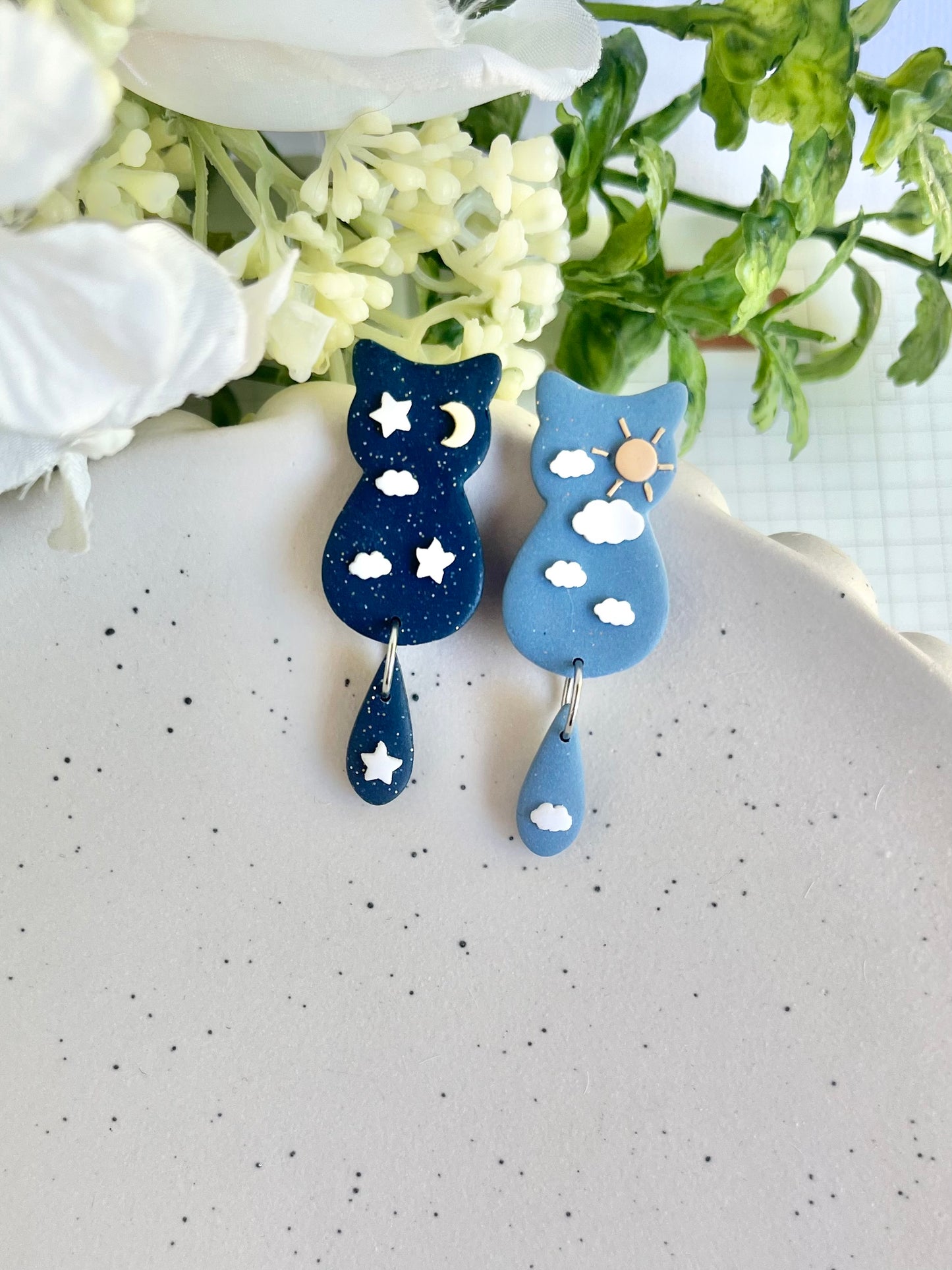 Blue cat-shaped earrings with star and moon designs on a white surface with flowers in the background.
