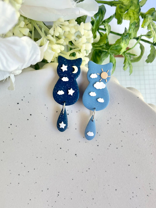 Blue cat-shaped earrings with star and moon designs on a white surface with flowers in the background.