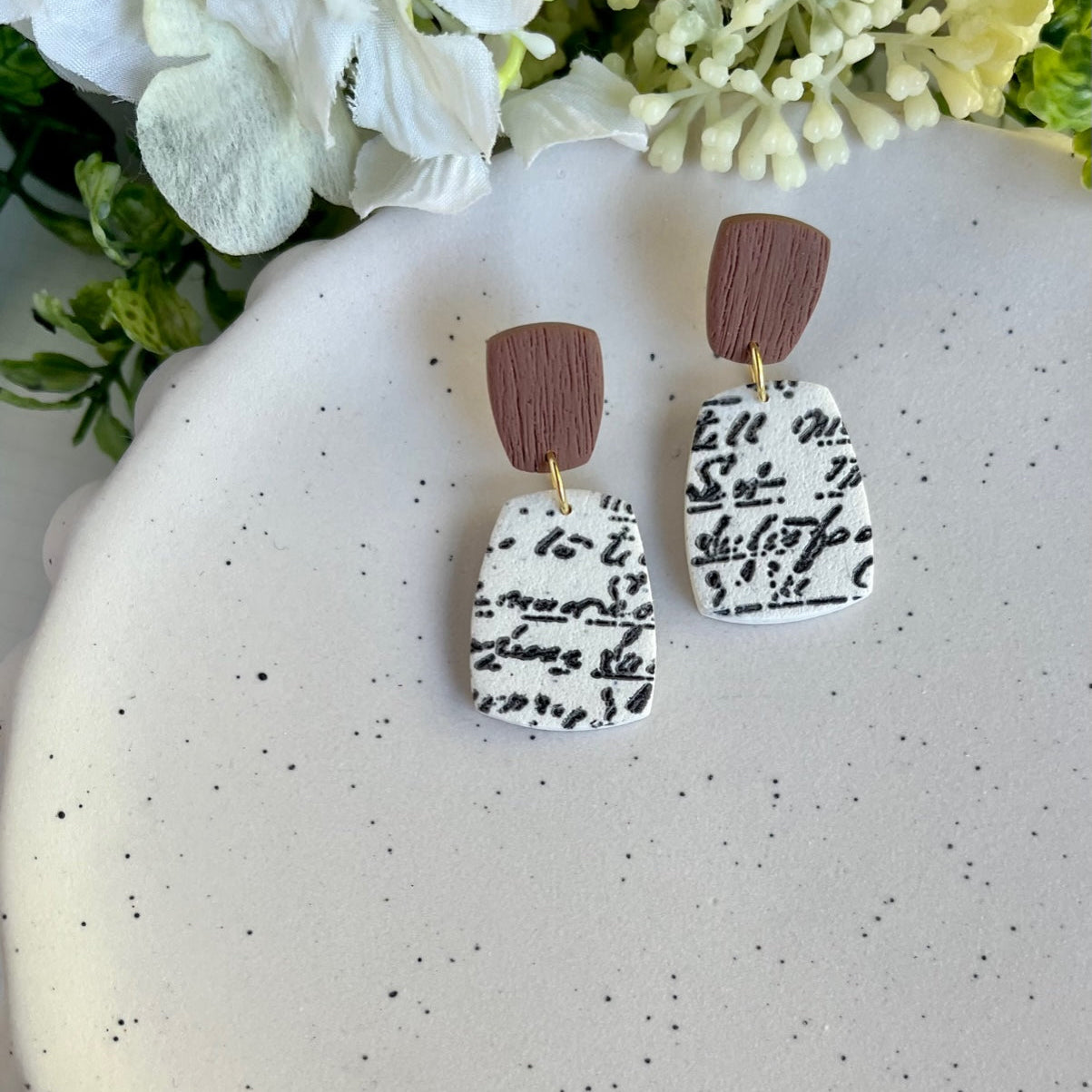 Pair of earrings with brown and black patterns on a speckled surface with flowers in the background