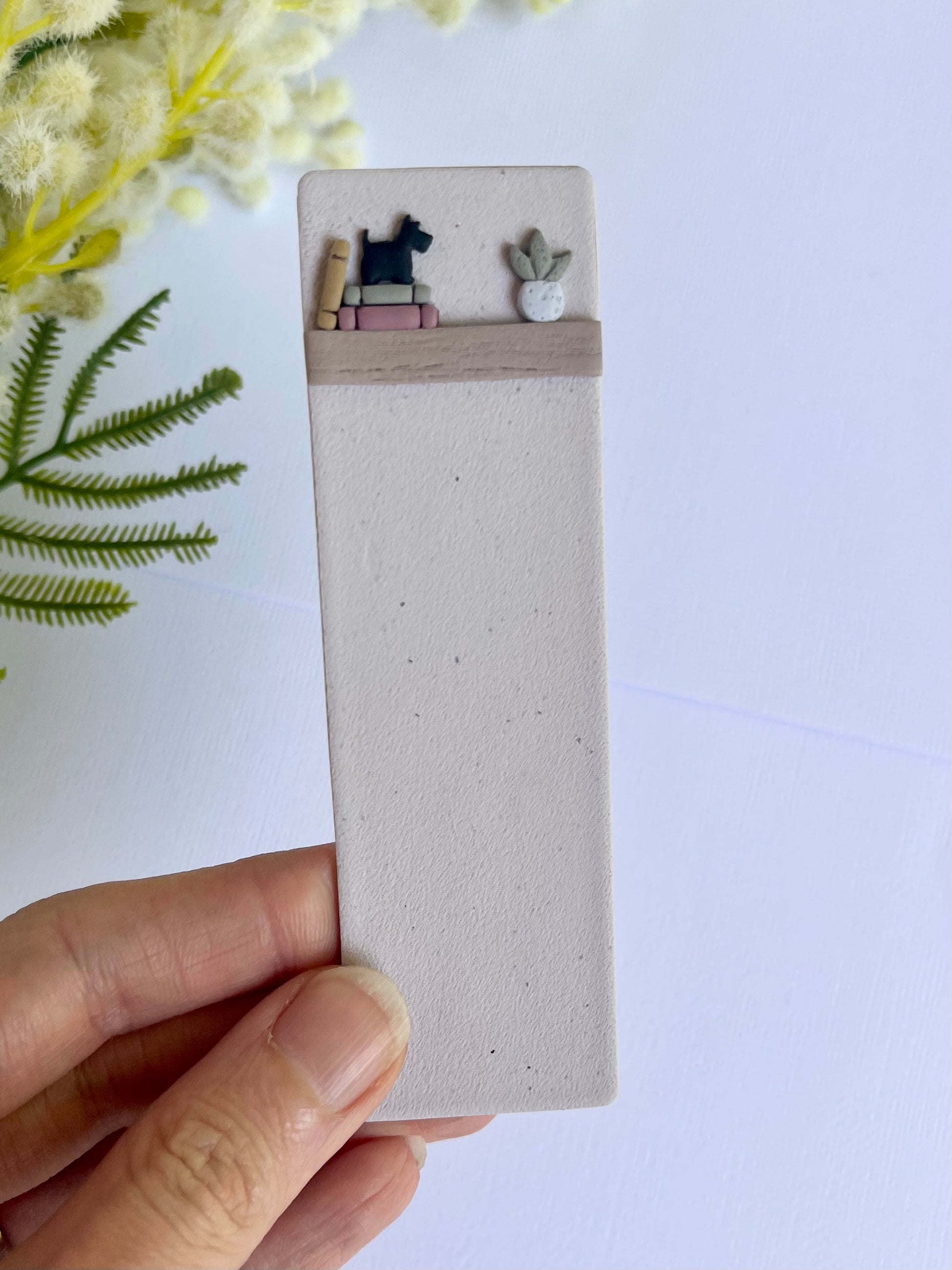 Hand holding a small rectangular object with miniature books and a cat figurine on a light background.