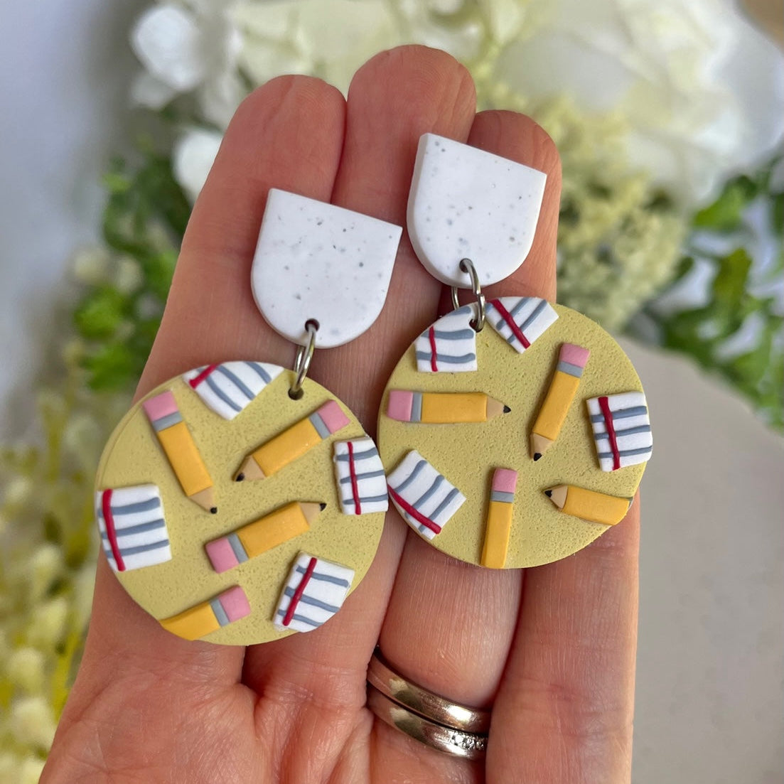 Hand holding a pair of earrings with pencil and book designs against a blurred floral background