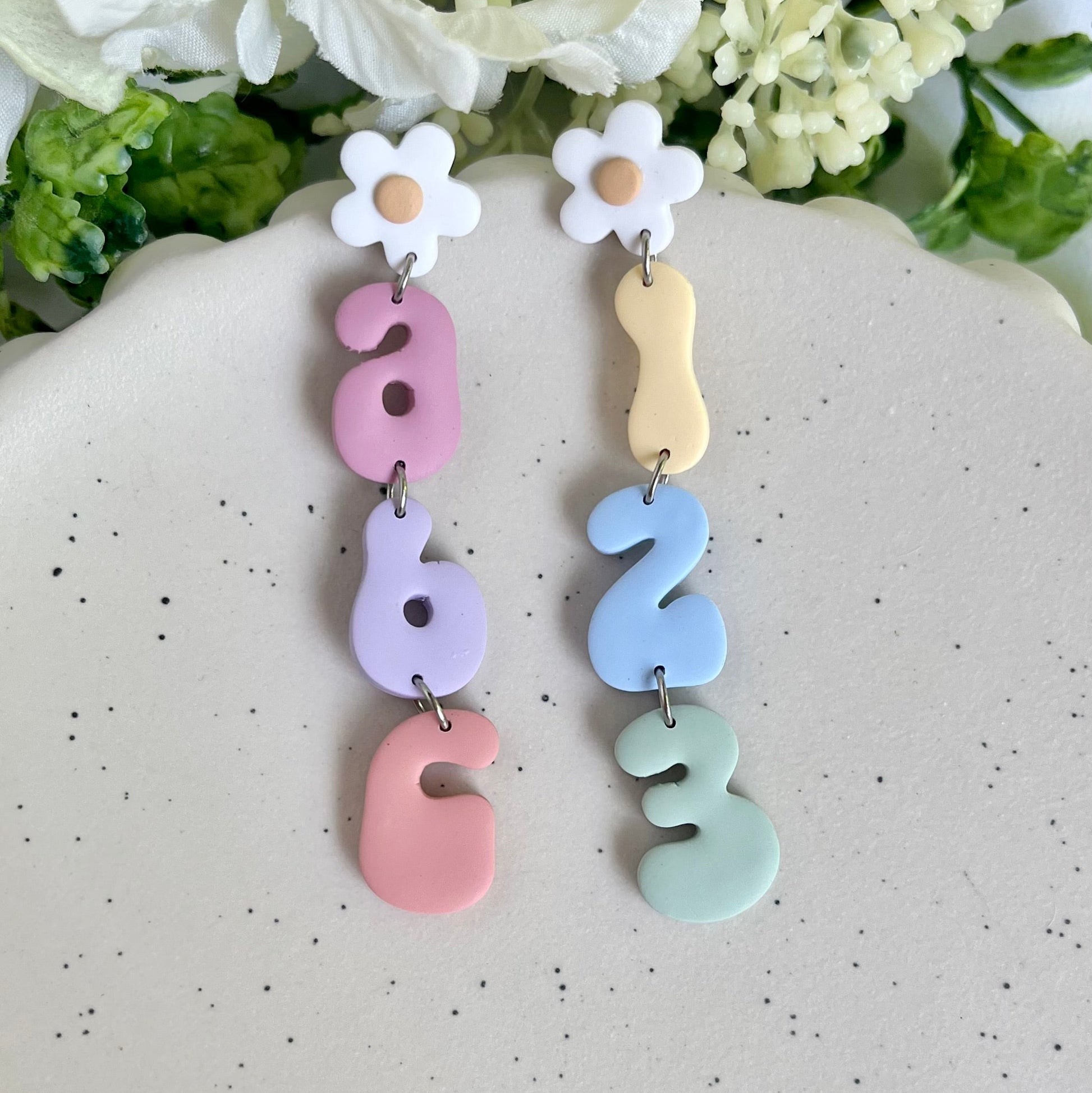 Pastel-colored number earrings with flowers on a speckled surface with flowers in the background