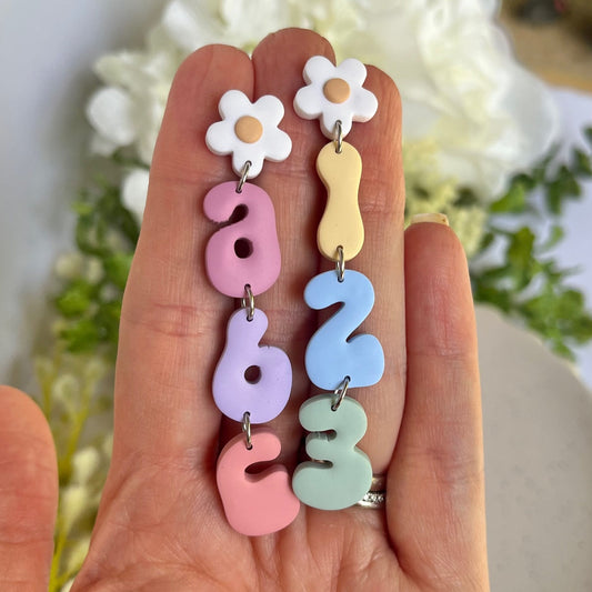 Colorful number and flower-shaped earrings held in a hand with a floral background.
