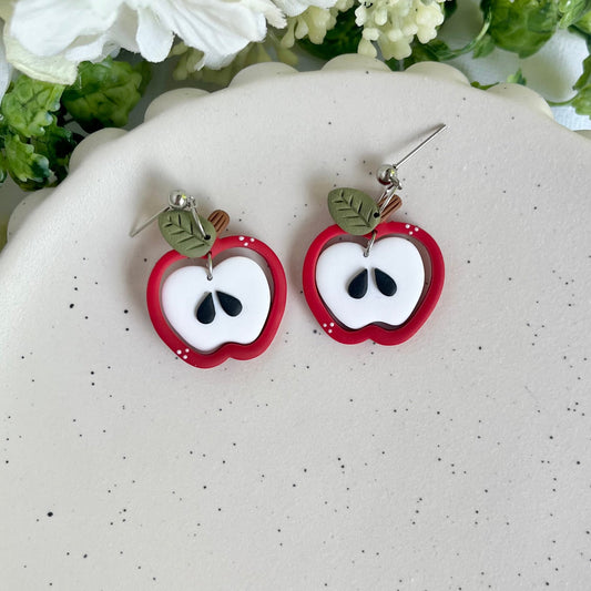 Apple-shaped earrings on a white surface with flowers in the background