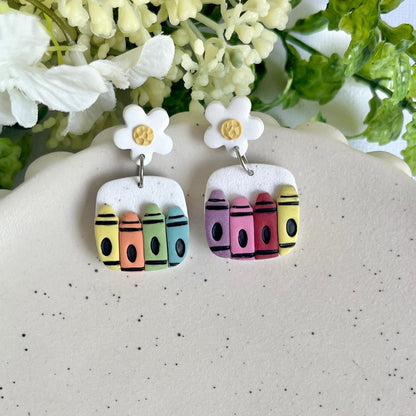 Crayon-shaped earrings on a white surface with flowers in the background
