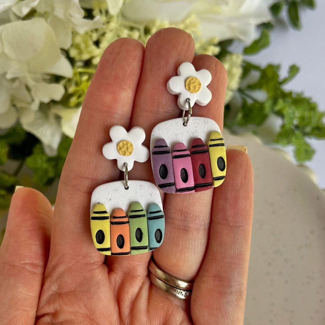 Colorful crayon-shaped earrings with flowers held in a hand against a floral background