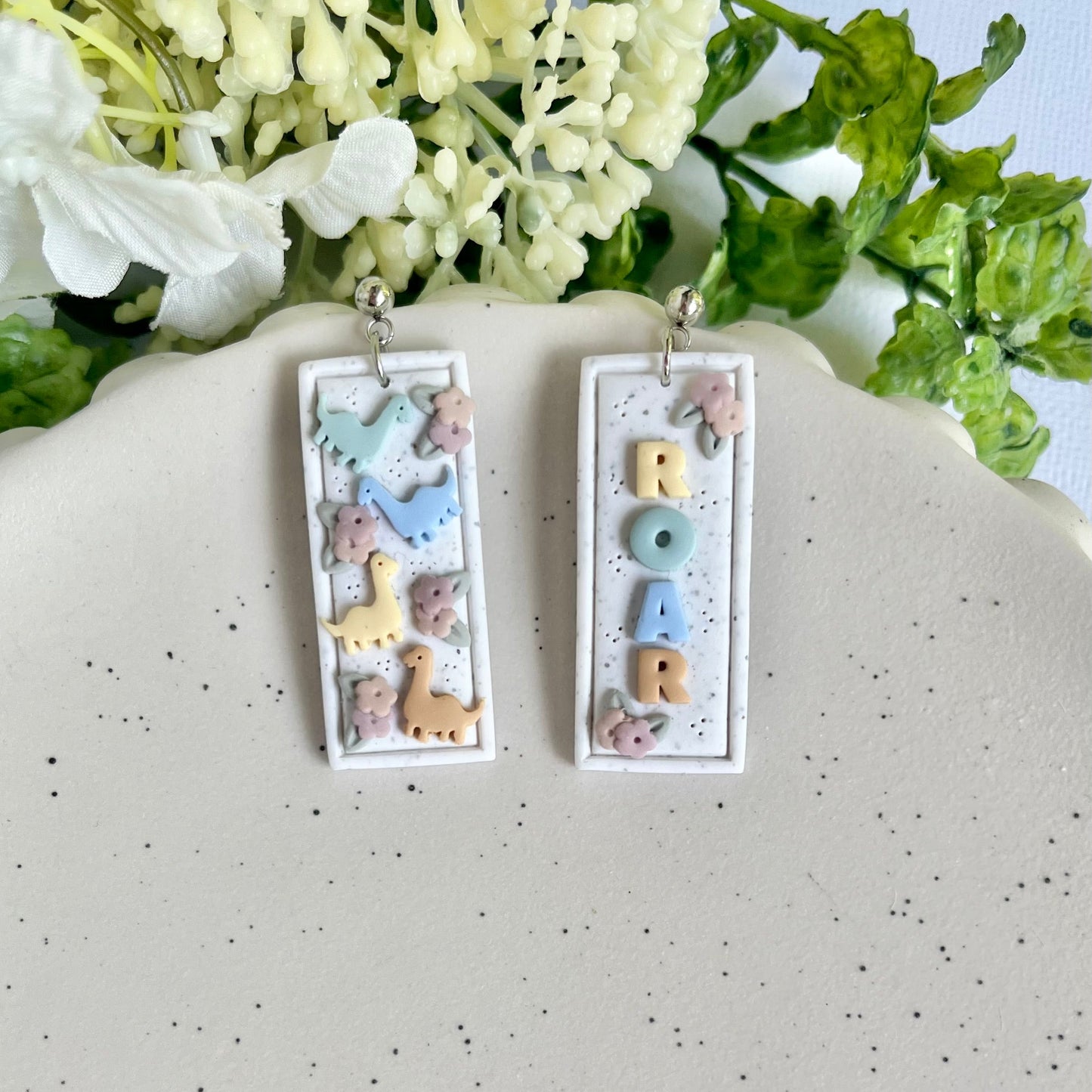 Pair of earrings with letter 'M' design on a speckled surface with flowers in the background