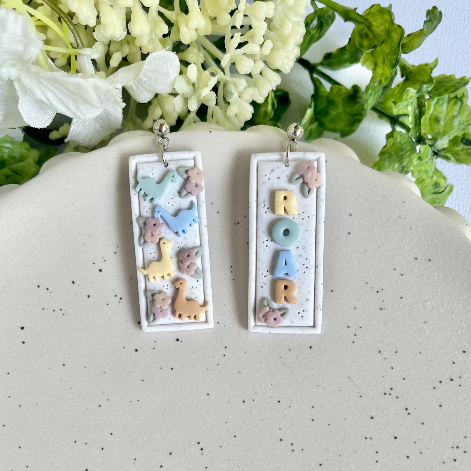 Pair of earrings with letter 'M' design on a speckled surface with flowers in the background