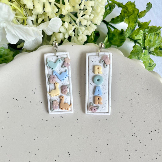 Pair of earrings with letter 'M' design on a speckled surface with flowers in the background