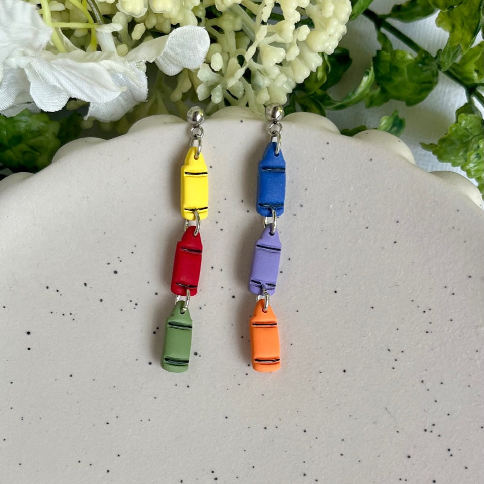 Colorful crayon-shaped earrings on a white surface with greenery in the background