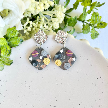 Pair of earrings with space-themed design on a white surface with greenery.