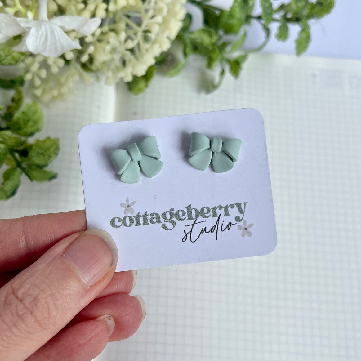 Pair of light blue bow-shaped earrings on a 'Cottageberry Studio' card held by a hand with a floral background.