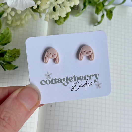 Pink bunny-shaped earrings on a card with 'cottageberry studio' branding, held over a checkered background.