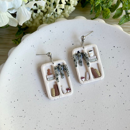 A pair of drop earrings with white, gold, and multicolor details designed to resemble bookshelves, displayed on a plate.