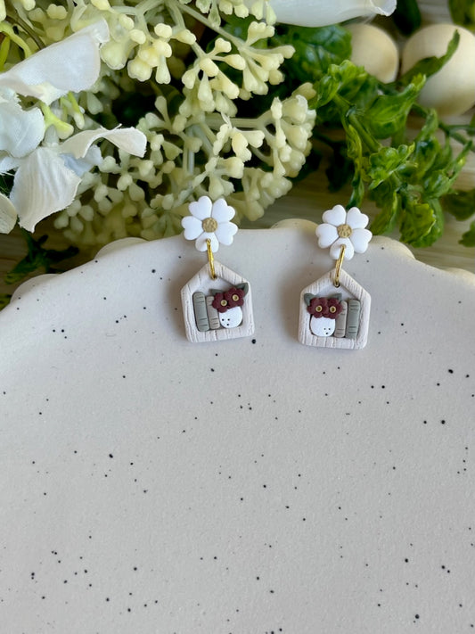 A pair of stud earrings designed to look like miniature bookshelves with white flowers and books, placed on a ceramic dish with floral decor.