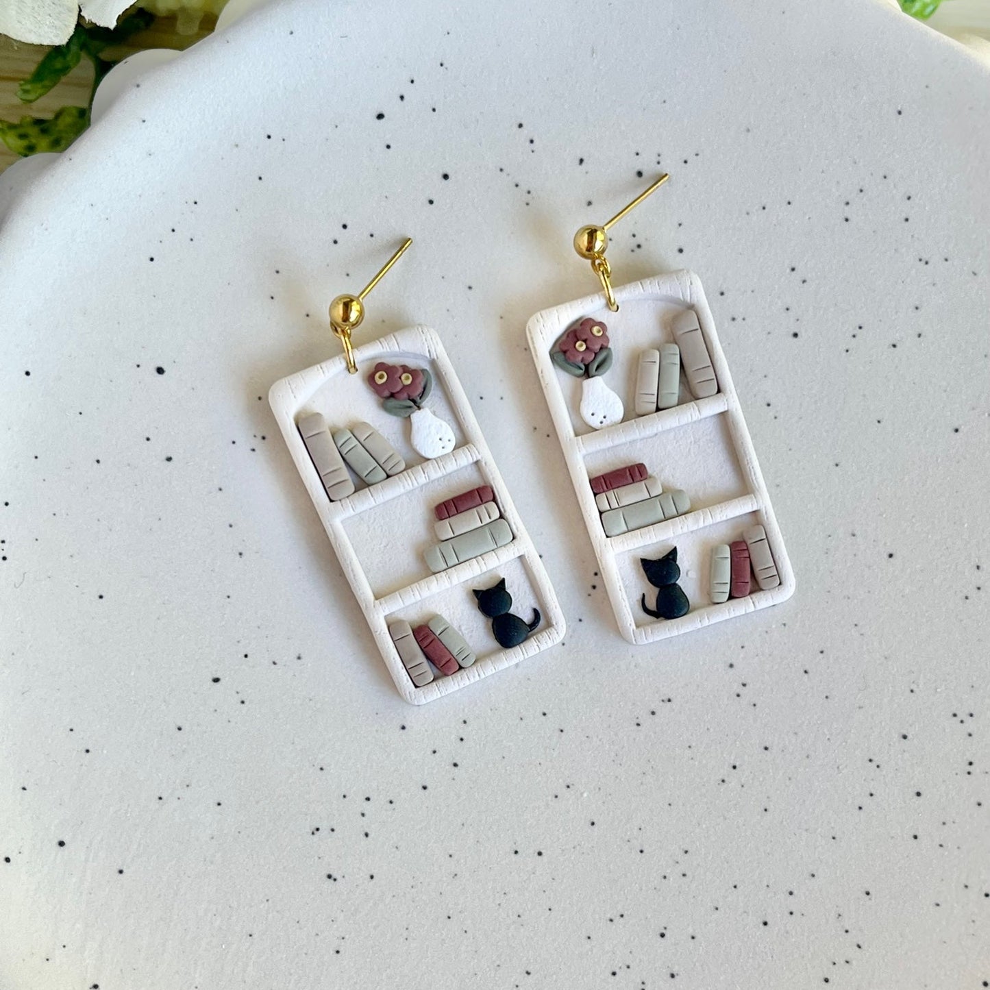 A pair of gold-colored bookshelf earrings adorned with miniature figures including a black cat and a flower pot, displayed on a white plate.