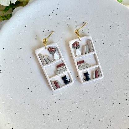 A pair of gold-colored bookshelf earrings adorned with miniature figures including a black cat and a flower pot, displayed on a white plate.