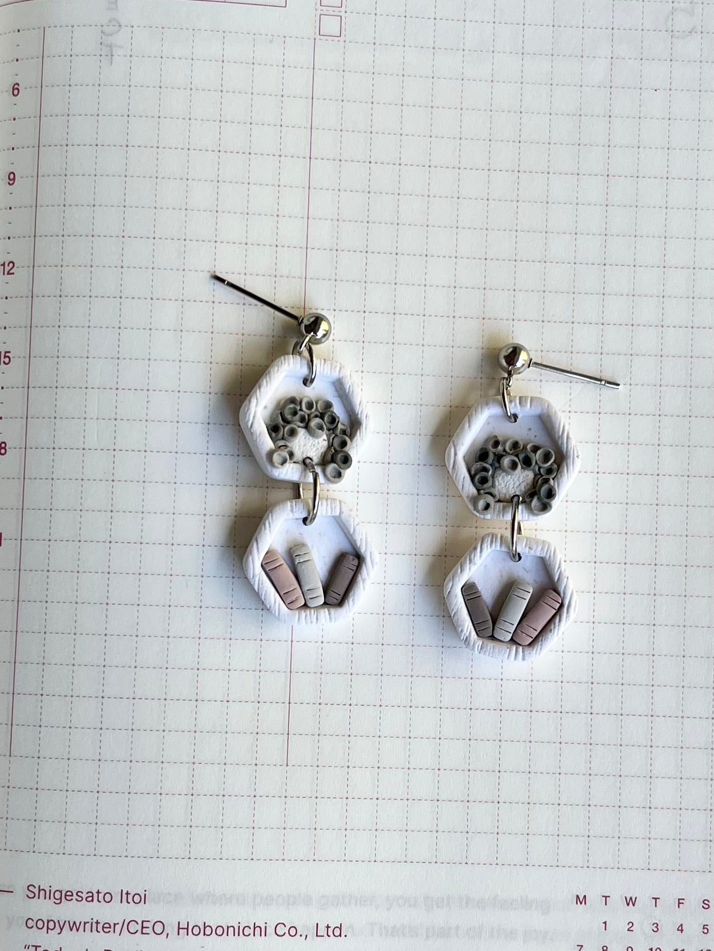 A pair of white hexagon-shaped earrings with a black plant design and miniature books, displayed on a grid paper background.