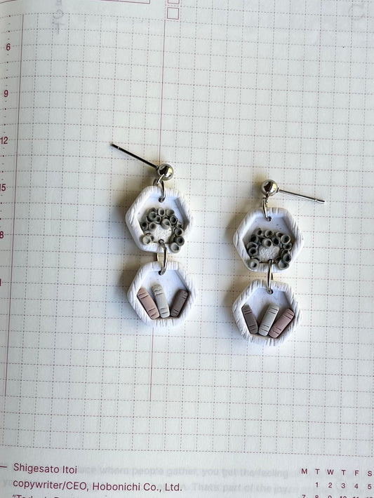 A pair of white hexagon-shaped earrings with a black plant design and miniature books, displayed on a grid paper background.