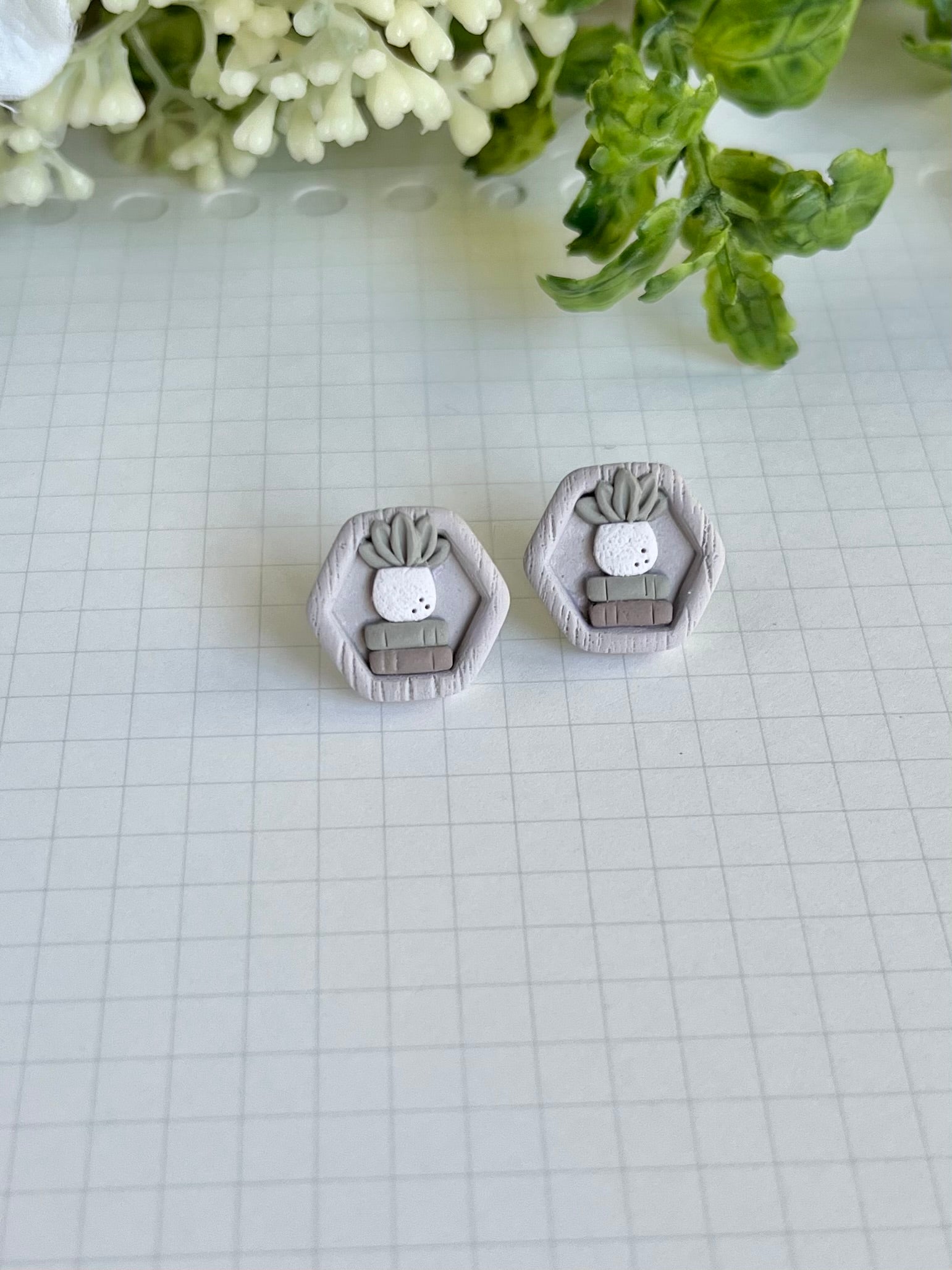 A pair of stud earrings designed to look like a miniature bookshelf with a potted plant, made from white material on a checkered background.